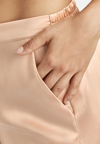 Pantalons en satin pêche clair avec une taille élastique, comportant une poche latérale où une main est posée. Texture lisse et design élégant.