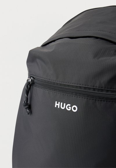 Gros plan sur un sac à dos HUGO noir, avec son rabat supérieur incurvé, sa poche avant zippée et son logo blanc.