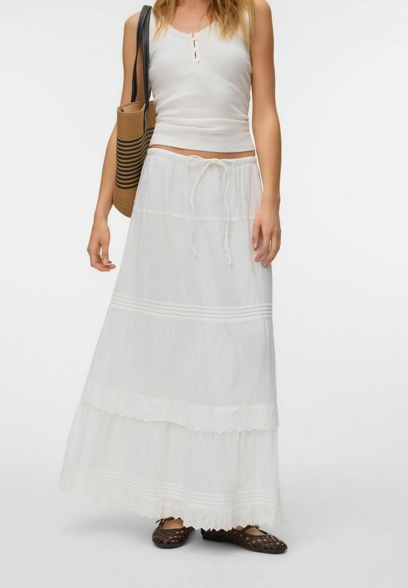 Femme portant un haut blanc sans manches, une longue jupe blanche avec cordon, des ballerines tressées marron et portant un sac fourre-tout rayé marron et noir.