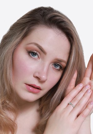 Nahaufnahme eines Gesichts mit heller Haut und langen, hellbraunen Haaren, das einen silbernen Ring mit kleinen, klaren Edelsteinen an einer Hand trägt.
