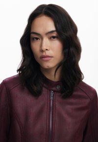 Maroon leather jacket with a high collar, zip closure, and textured finish. Features subtle stitching details and a sleek design.