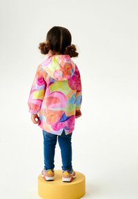 Veste imperméable colorée et légère avec une capuche, arborant un motif abstrait vibrant en rose, orange et bleu. Associée à un jean bleu et des baskets.