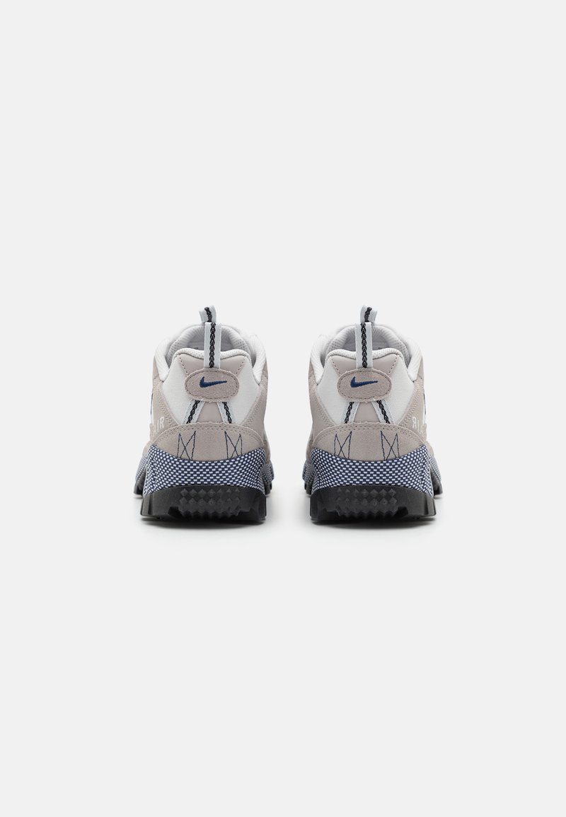 Baskets de sport grises et blanches avec une tige en mesh texturé et en daim, accents navy et semelle noir robuste. Comprend un col rembourré et des poignées.