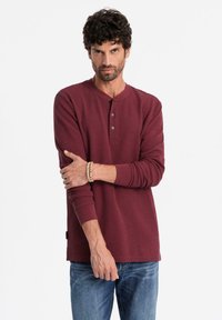 Uomo con capelli ricci e scuri che indossa una maglietta henley a maniche lunghe color mogano e jeans blu, mentre sistema la manica della maglietta con braccialetti di legno.