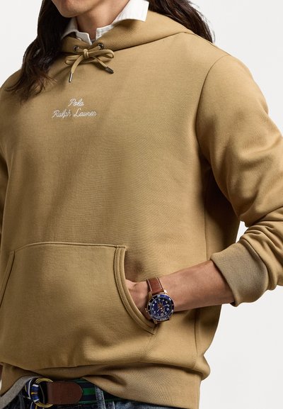 Μπεζ φούτερ με κορδόνι, μπροστινή τσέπη και λογότυπο. Ο ιστός φαίνεται μαλακός, συμπληρωμένος από ένα μπλε και χρυσό ρολόι με δερμάτινο λουρί.