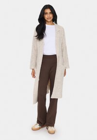 Kvinna klädd i en lång beige öppen cardigan, vit t-shirt, bruna vida byxor och beige sneakers, stående mot en enfärgad bakgrund.
