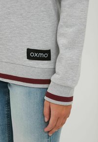 Grå hettegenser med maroonstripete mansjetter og nederkant. Har en svart etikett med merkenavnet 'OXMO'. Myk stoff, avslappet design.