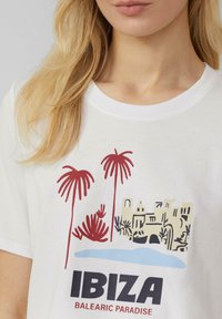 T-shirt blanche avec une impression graphique de palmiers, d'un château et d'eau, avec le texte "IBIZA BALEARIC PARADISE" en noir et rouge.