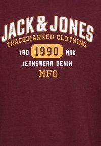 Kastanienbraunes Baumwoll-T-Shirt mit weißen und gelben Schriftzügen: "JACK & JONES," "TRADEMARKED CLOTHING," und "1990." Einfaches Grafikdesign mit Rundhalsausschnitt.
