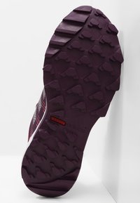 Suela de zapato deportivo morado, con una superficie de goma texturizada con tacos profundos, lados reforzados y un acento de logo rojo.