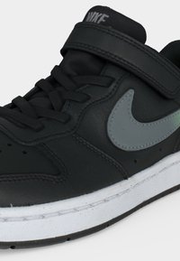 Μαύρο αθλητικό παπούτσι Nike με γκρι λογότυπο swoosh, μαύρα κορδόνια, λουράκι με βέλκρο και λευκή σόλα σε απλό λευκό φόντο.