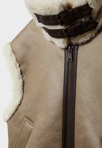 Beige vesta z beránka s vysokým límcem, hnědými koženými pásky a přezkami, a předním zipem podél švu pro zapínání.