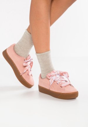 Nogi w jasnobeżowych skarpetkach i jasnoróżowych tenisówkach Puma z grubymi brązowymi podeszwami i markowymi wstążkowymi sznurowadłami na białym tle.