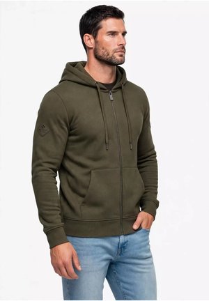 Un uomo che indossa una felpa verde oliva con zip e tasche frontali e jeans azzurri, in piedi con una mano in tasca, guarda verso destra.