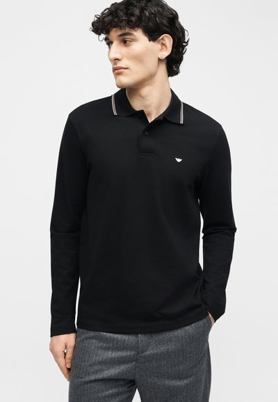 Νεαρός με σγουρά σκούρα μαλλιά που φοράει μαύρο πόλο με μακριά μανίκια και ριγέ γιακά, καθώς και γκρι παντελόνι, κοιτάει προς τα αριστερά.