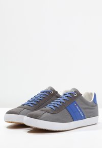 Baskets en toile grise avec accents bleus, semelle en caoutchouc blanche et lacets bleus. Comprend une bande logo latérale et un bout arrondi.