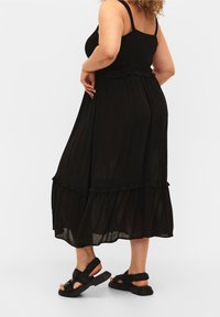 Vestido negro sin mangas con un corpiño fruncido y un dobladillo con volantes, hecho de tela ligera. Se lleva con sandalias negras con tiras y una suela gruesa.