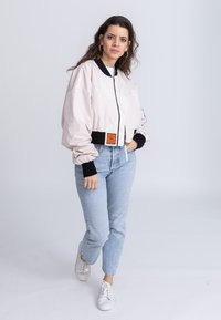 Helle pink Bomberjacke mit schwarzen Akzenten und einem Reißverschluss vorne. Kombiniert mit hellblauen Jeans und weißen Sneakers. Verfügt über ein oranges Etikett.
