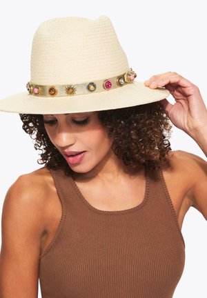 Vrouw met krullend haar die een beige hoed met brede rand draagt, versierd met een kleurrijke band met juwelen, en die de rand van de hoed met haar rechterhand aanpast, gekleed in een bruin mouwloos topje.