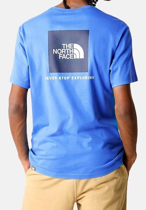 Mężczyzna noszący niebieską koszulkę The North Face z logo i tekstem "Never Stop Exploring" na plecach, w połączeniu z beżowymi spodniami.