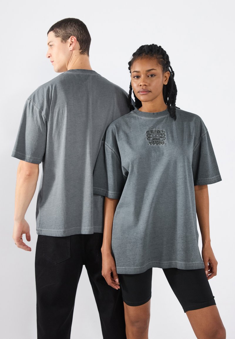 Graue übergroße T-Shirts aus weichem Material mit kurzen Ärmeln und einem mittig platzierten Grafikdruck. Modell trägt Shorts und dunkle Hosen.