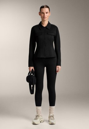 Nő áll, fekete, testhezálló dzsekit és leggingset visel, kis fekete táskát tart a kezében, világos bézs zoknival és fehér tornacipővel.