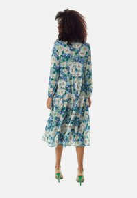 Floral midi dress in blue and green hues with long sleeves, gathered waist, tiered skirt, and delicate print. Worn with green heeled sandals.