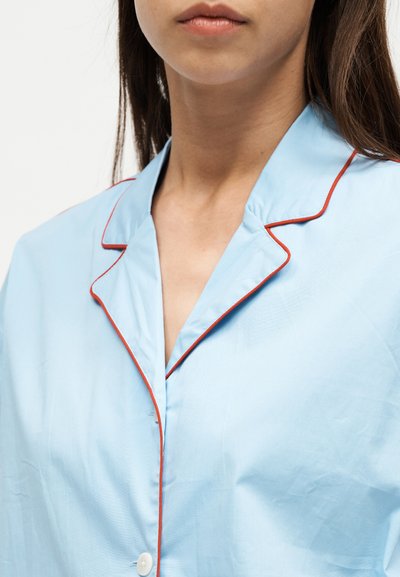 Femme portant une chemise bleu clair boutonnée avec un liseré rouge le long du col et du bord avant, visible du cou à la poitrine.
