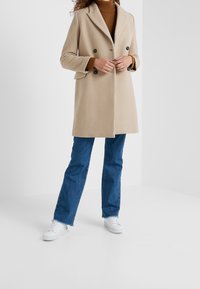 Beige Wolle-Mix Mantel mit doppelter Knopfleiste und Reverskragen, seitlichen Taschen und schwarzen Knöpfen, kombiniert mit blauen geraden Jeans und weißen Sneakers.