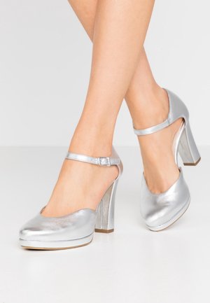 Jambes de femme portant des chaussures Mary Jane à talons blocs métalliques argentés avec brides à la cheville sur un sol blanc.