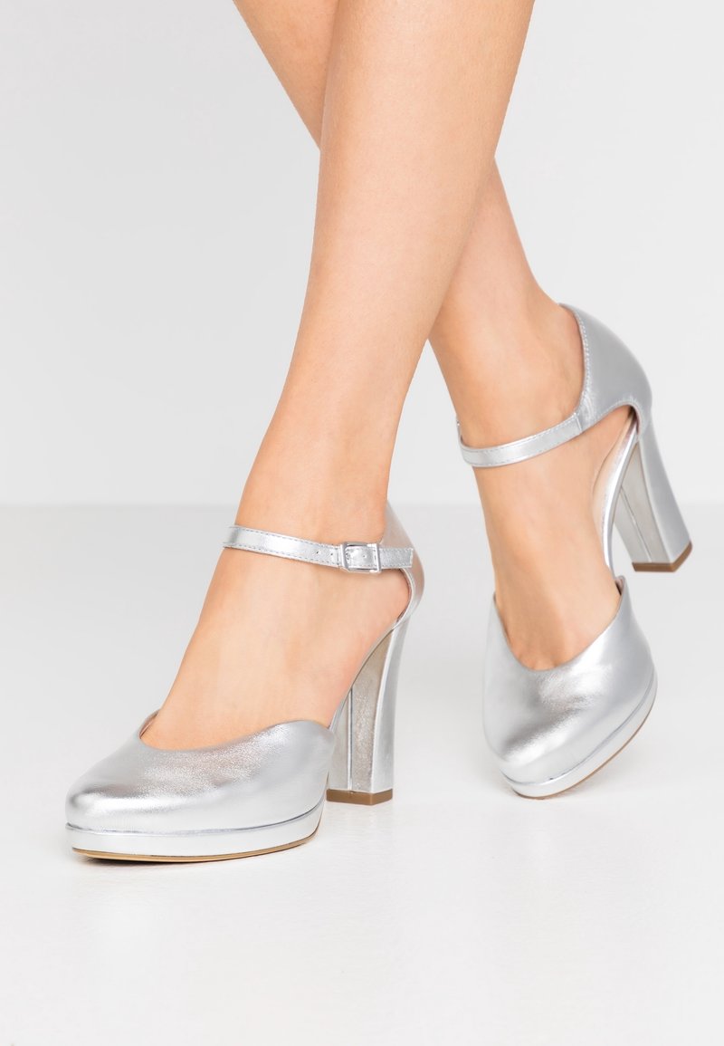 Jambes de femme portant des chaussures Mary Jane à talons blocs métalliques argentés avec brides à la cheville sur un sol blanc.