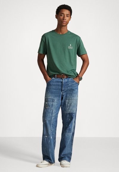 Green cotton t-shirt with a small logo, paired with blue distressed, paint-splattered denim pants featuring multiple pockets. White sneakers.