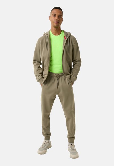 Khaki trainingspak bestaande uit een sweatshirt met capuchon en joggingbroek, in combinatie met een felgroen t-shirt. De schoenen zijn lichtgrijs met chunky zolen.