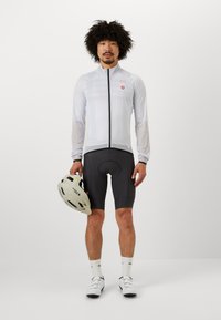 Veste de cyclisme légère blanche avec une fermeture éclair, shorts rembourrés noirs, chaussures de cyclisme blanches et un casque gris clair, tenu dans la main gauche.