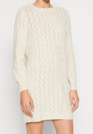 Kvinde iført en cremefarvet, langærmet strikkjole med zigzag-mønster, ribbet manchetter og ribbet kant.