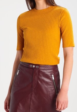 Mosterdgele ribbelcrop top met korte mouwen, gecombineerd met een bordeauxleren rok met zilverkleurige ritszakken en drukknopdetails.