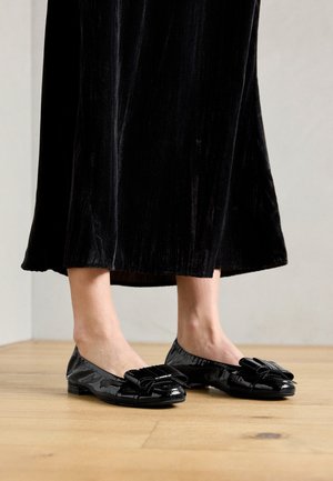 Zwarte gepatineerde ballerina's met een strikaccent op de teen. Gladde textuur, ronde teen en een lage platte zool. Set gedragen met een zwarte fluwelen rok.