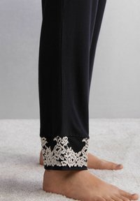 Zwarte broek met een geweven kantmanchet met witte bloemenpatronen. Glad stofafwerking, aansluitende pasvorm, enkel lengte en blote voeten zichtbaar.