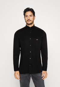 Homme aux cheveux bouclés portant une chemise noire boutonnée et un jean gris, debout devant un fond clair uni.