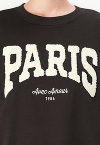 Brown sweatshirt featuring large cream textured letters reading "PARIS" and smaller white text "Avec Amour 1986" below.