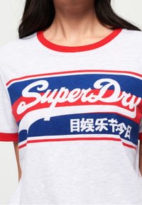 Graues T-Shirt mit roten und blauen Streifenakzenten, das auffälligen weißen "SuperDry"-Text und asiatische Zeichen auf der Brust zeigt.