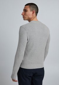 Bărbat îmbrăcat cu un pulover texturat în gri deschis cu mânecă lungă și pantaloni închiși, întors ușor, privind spre stânga, pe un fundal simplu.