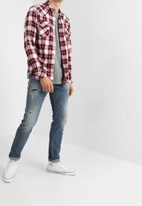 Röd och svart rutigt flanellskjorta med två bröstfickor, grå t-shirt under, slitna blå jeans och vita sneakers.