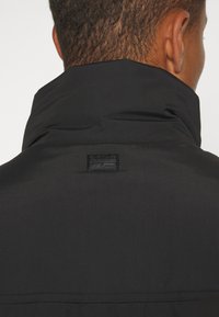 Närbild av ryggen på en person som bär en svart jacka med hög krage och en liten rektangulär logotyp som är centrerad under kragen.