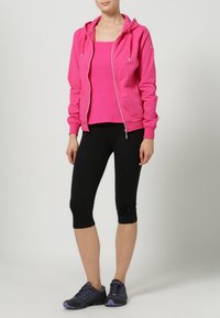 Vaaleanpunainen vetoketjullinen huppari, vaaleanpunainen toppi, mustat capri-legginssit ja mustat urheilukengät. Sileä kangas, istuva malli, nyörivivahtein.