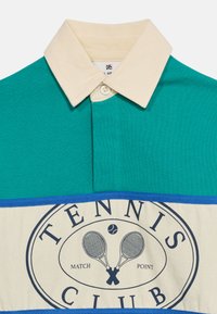 Πόλο με γιακά σε τιρκουάζ και κρεμ χρώμα με μπλε περιθώριο, με κείμενο "Tennis Club" και λογότυπο με σταυρωτές ρακέτες τένις και μπάλα τένις.