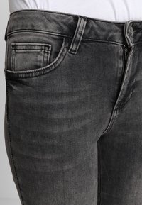 Jeans de mezclilla negra con un acabado desgastado, que cuentan con un diseño de cinco bolsillos, presillas para el cinturón y cierre frontal con cremallera. Tejido texturizado con detalles de costura sutiles.