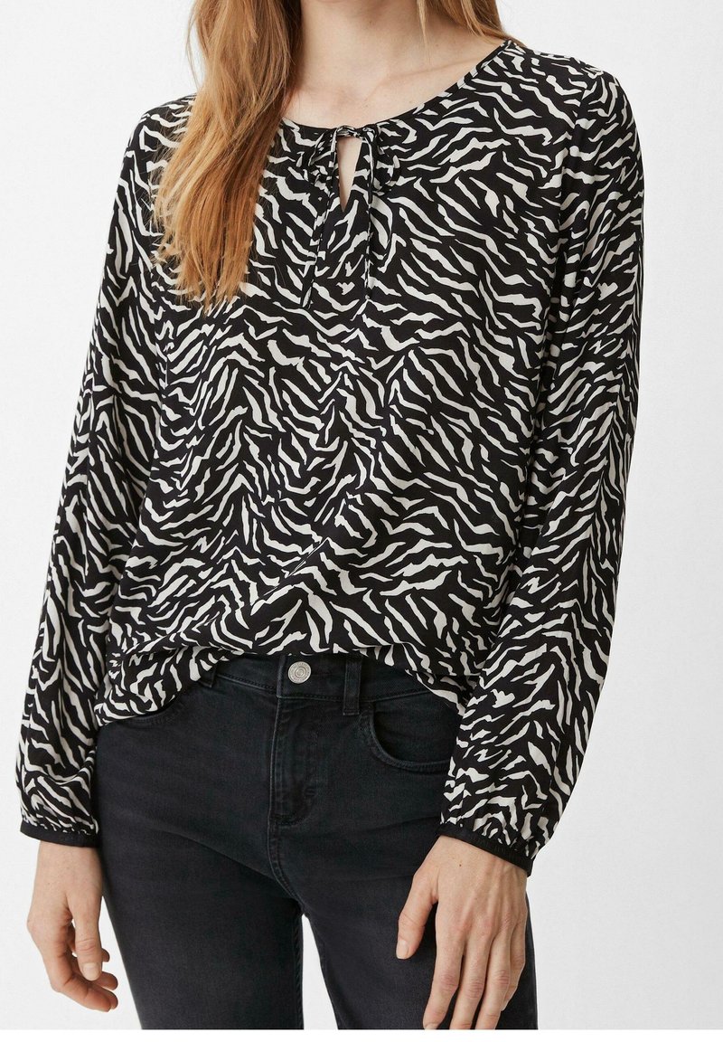 Femme portant un chemisier à manches longues imprimé zèbre noir et blanc avec une encolure en forme de trou de serrure et un pantalon noir.