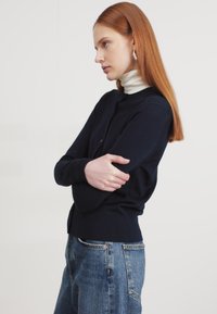 Mornarji jopič z okroglim ovratnikom, dolgimi rokavi in gumbi, v kombinaciji z belim puloverjem s turtleneckom in modrimi kavbojkami. Gladka tekstura.