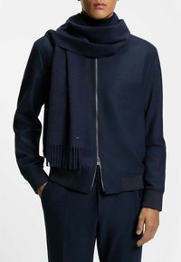 Sciarpa di lana blu navy con frange, drappeggiata attorno al collo, abbinata a una giacca blu navy con zip e polsini e orlo a costine.